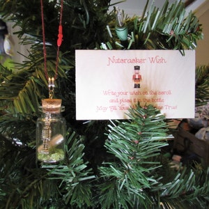 May include: A glass bottle ornament with a cork stopper and a silver nutcracker figurine inside. The bottle is filled with green glitter and a rolled up piece of paper. A card with the text "Nutcracker Wish" is attached to the bottle. The card also includes instructions to write a wish on the paper and place it in the bottle.