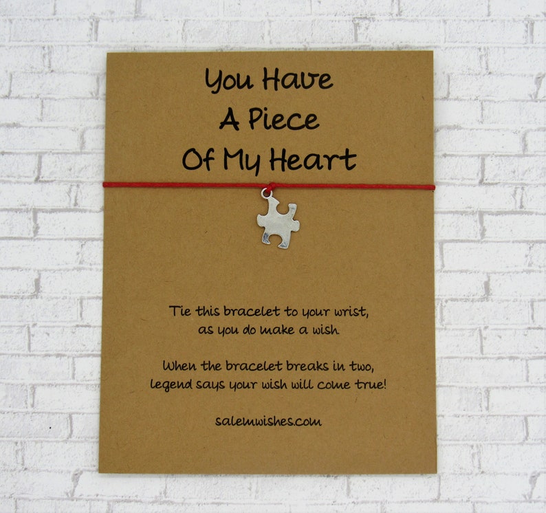 K&ouml;nnte beinhalten: Eine braune Karte mit dem Text "You Have A Piece Of My Heart" und einer roten Schnur mit einem silbernen Puzzle-St&uuml;ck-Anh&auml;nger. Der Text unter der Karte lautet "Binde dieses Armband an dein Handgelenk, w&auml;hrend du einen Wunsch machst. Wenn das Armband in zwei Teile zerbricht, besagt die Legende, dass dein Wunsch in Erf&uuml;llung geht! salemwishes.com"