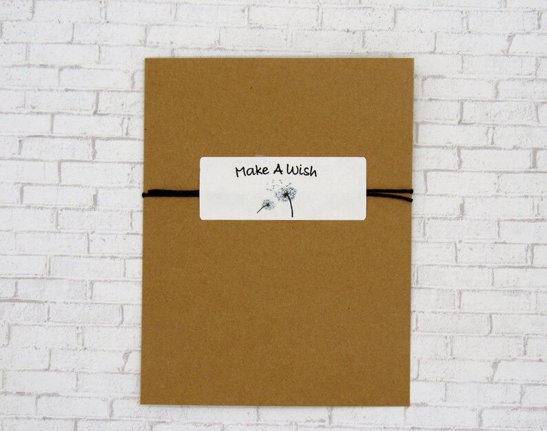 May include: A brown card with a white label that reads "Make A Wish" in black script. The label features a black and white dandelion illustration. A black string is wrapped around the card.