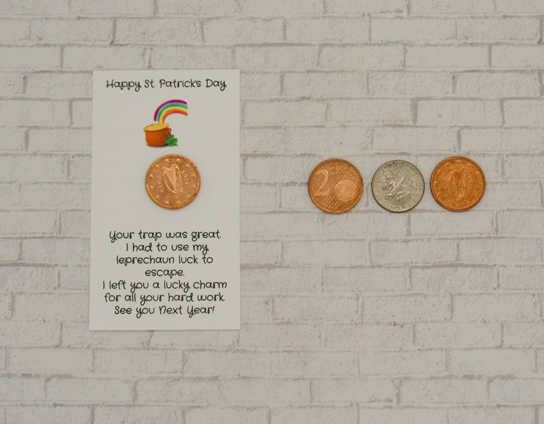 May include: A white card with a green shamrock and rainbow pot of gold illustration. The card reads "Happy St. Patrick's Day". The text on the card reads "Your trap was great. I had to use my leprechaun luck to escape. I left you a lucky charm for all your hard work. See you next year!" There are four Euro coins on a white brick background.