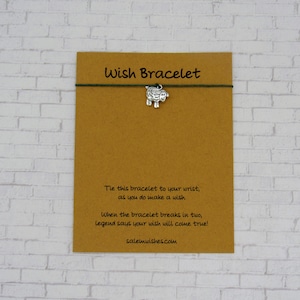 May include: A brown card with the text "Wish Bracelet" printed in black. The card has a green string tied around it with a silver charm of a sheep attached. The text on the card reads: "Tie this bracelet to your wrist, as you do make a wish. When the bracelet breaks in two, legend says your wish will come true! salemwishes.com"