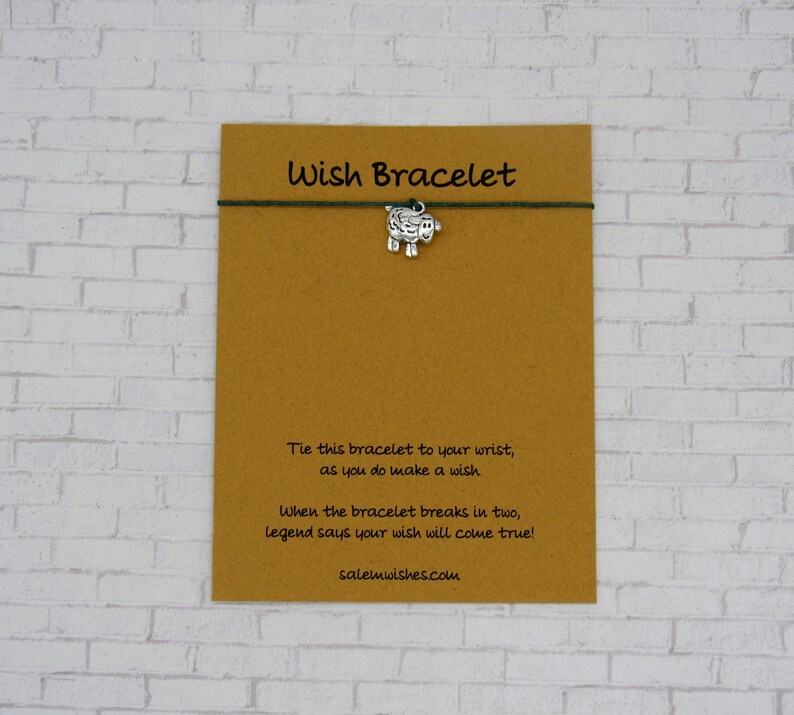 May include: A brown card with the text "Wish Bracelet" printed in black. The card has a green string tied around it with a silver charm of a sheep attached. The text on the card reads: "Tie this bracelet to your wrist, as you do make a wish. When the bracelet breaks in two, legend says your wish will come true! salemwishes.com"