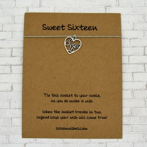May include: A brown card with the text "Sweet Sixteen" and a silver heart-shaped charm with the number "16". The card also includes text about making a wish. The charm is attached to the card with a thin, light green string.