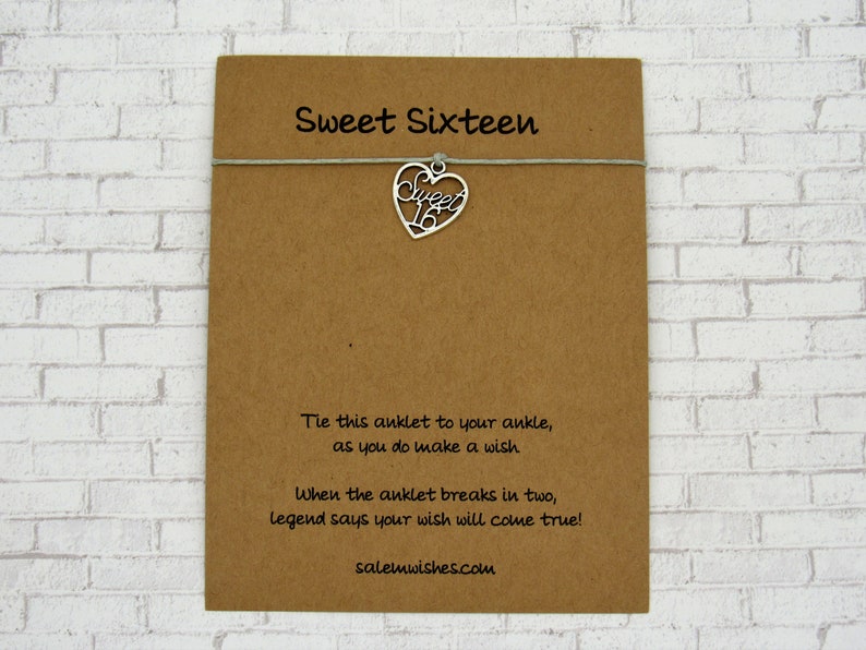May include: A brown card with the text "Sweet Sixteen" and a silver heart-shaped charm with the number "16". The card also includes text about making a wish. The charm is attached to the card with a thin, light green string.