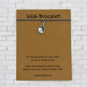 May include: A brown card with the text "Wish Bracelet" printed in black. A blue string with a silver charm of a bagpipe is attached to the card. The text on the card reads: "Tie this bracelet to your wrist, as you do make a wish. When the bracelet breaks in two, legend says your wish will come true! salemwishes.com"