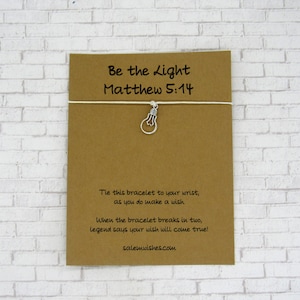 May include: A brown card with the text "Be the Light Matthew 5:14" and a small silver lightbulb charm attached to a white string. The card also includes the text "Tie this bracelet to your wrist, as you do make a wish. When the bracelet breaks in two, legend says your wish will come true! salemwishes.com".