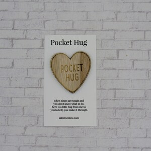May include: A white card with the text "Pocket Hug" printed on it. A small wooden heart with the text "Pocket Hug" is attached to the card. The card has a message about sending a hug when times are tough.