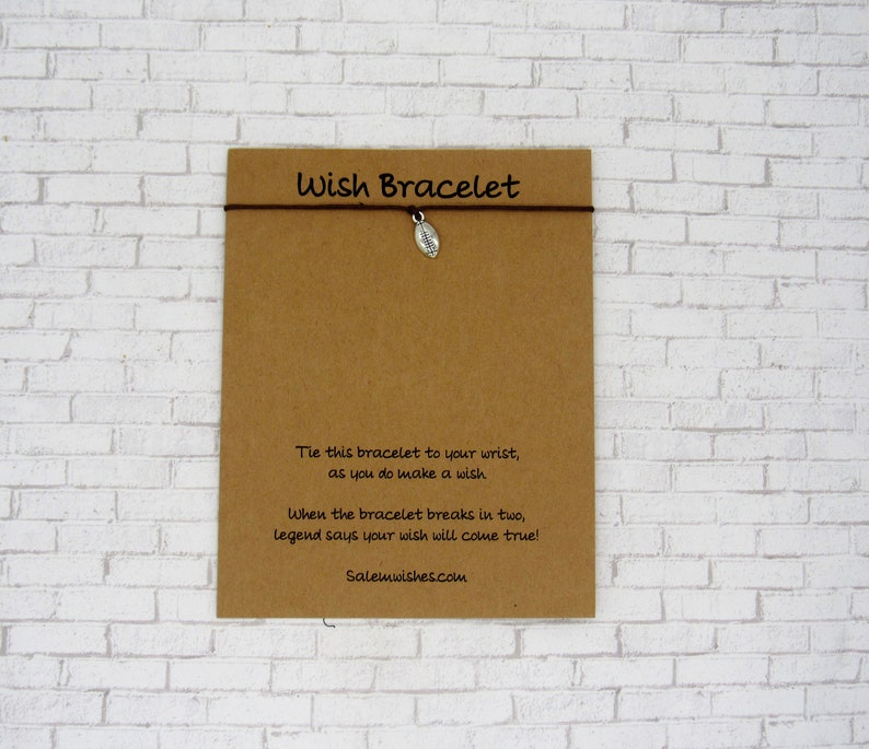 May include: A brown string bracelet with a small silver-toned football charm attached to a rectangular kraft paper card. The card features the text Wish Bracelet and a poem about making a wish, along with the website address Salemwishes.com.