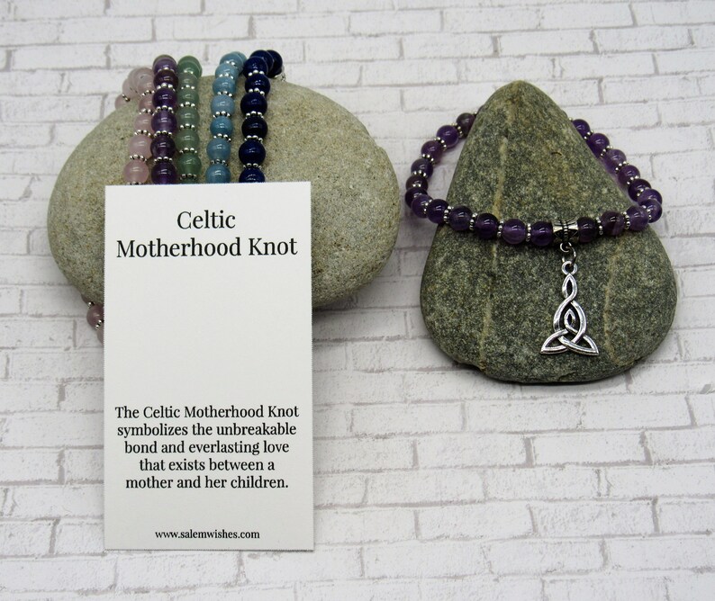K&ouml;nnte beinhalten: Ein violettes Amethyst-Perlenarmband mit einem silbernen keltischen Knoten-Anh&auml;nger. Das Armband liegt auf einem grauen Stein mit einer wei&szlig;en Karte, auf der "Celtic Motherhood Knot" und "The Celtic Motherhood Knot symbolizes the unbreakable bond and everlasting love that exists between a mother and her children. www.salemwishes.com" steht.