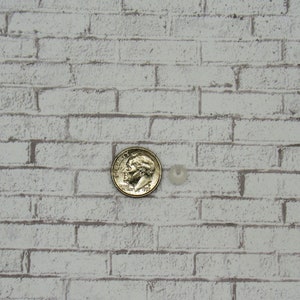 K&ouml;nnte beinhalten: Nahaufnahme einer wei&szlig;en Backsteinwand mit einer Silberm&uuml;nze und einer kleinen wei&szlig;en Perle in der Mitte des Bildes.