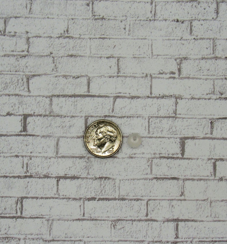 K&ouml;nnte beinhalten: Nahaufnahme einer wei&szlig;en Backsteinwand mit einer Silberm&uuml;nze und einer kleinen wei&szlig;en Perle in der Mitte des Bildes.