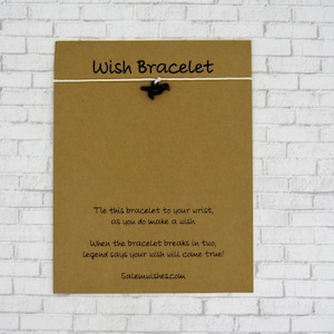 May include: A brown card with the text "Wish Bracelet" printed on it. The card has a white string tied to it with a black bird charm hanging from the string. The text on the card reads: "Tie this bracelet to your wrist, as you do make a wish. When the bracelet breaks in two, legend says your wish will come true! Salemwishes.com"