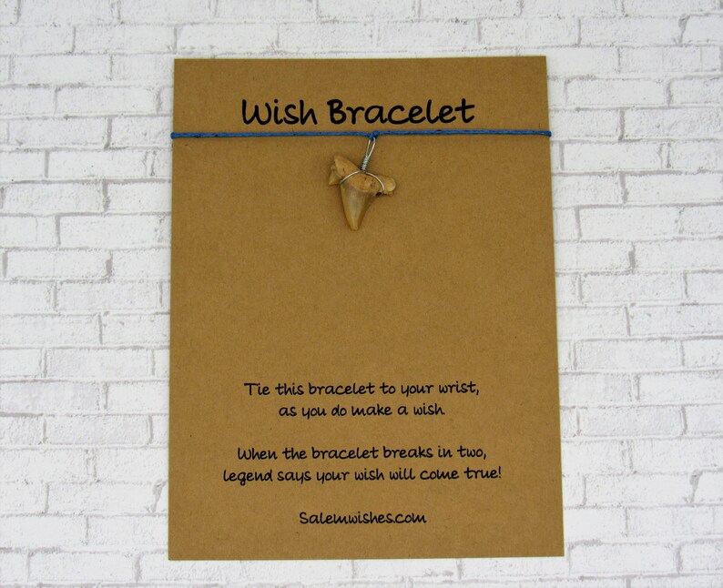 May include: A brown card with the text "Wish Bracelet" printed in blue. The card has a blue string with a fossilized shark tooth hanging from it. The text on the card reads: "Tie this bracelet to your wrist, as you do make a wish. When the bracelet breaks in two, legend says your wish will come true! Salemwishes.com"