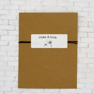 K&ouml;nnte beinhalten: Ein brauner Papierumschlag mit einem wei&szlig;en Etikett, auf dem "Make A Wish" steht und eine Schwarzwei&szlig;illustration von L&ouml;wenzahnpflanzen zu sehen ist. Der Umschlag ist mit einer schwarzen Schnur gebunden.