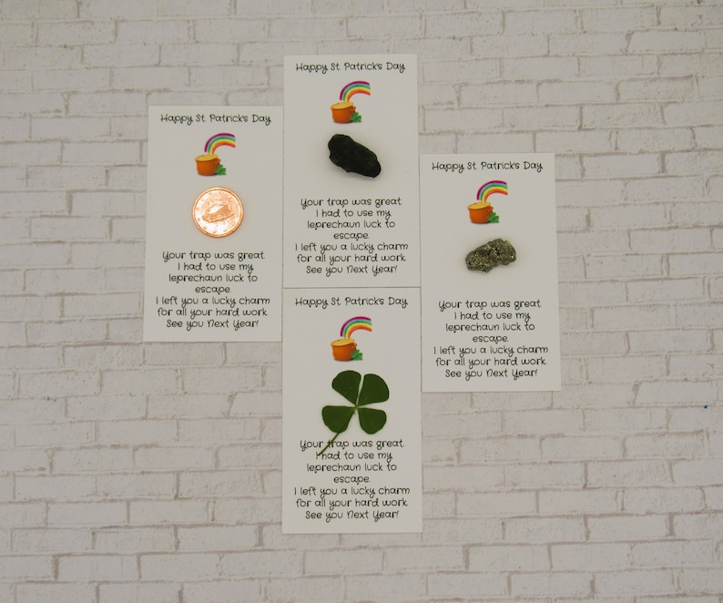 May include: Four white cards with a rainbow and pot of gold design. Each card has a different lucky charm attached: a coin, a black rock, a gold rock, and a four-leaf clover. The cards have the text "Happy St. Patricks Day" and a message about a leprechaun's luck.