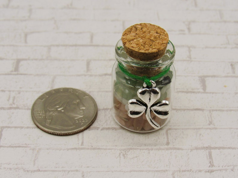 May include: A small glass jar filled with green, pink, and white stones. The jar has a cork lid and a silver shamrock charm hanging from it with a green ribbon.