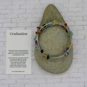 May include: A silver bracelet with a variety of colorful crystals, including red, orange, yellow, green, blue, and purple. The bracelet is on a gray rock with a card that says "Graduation" and lists the crystals and their meanings.