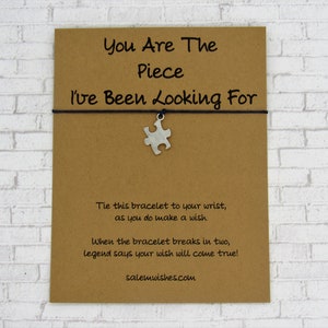 K&ouml;nnte beinhalten: Eine braune Karte mit dem Text "You Are The Piece I've Been Looking For" und einem silbernen Puzzle-St&uuml;ck-Anh&auml;nger, der an einer schwarzen Schnur befestigt ist. Die Karte enth&auml;lt auch Anweisungen f&uuml;r ein Gl&uuml;cksarmband: "Binde dieses Armband an dein Handgelenk, w&auml;hrend du einen Wunsch machst. Wenn das Armband in zwei Teile zerbricht, besagt die Legende, dass dein Wunsch in Erf&uuml;llung geht!" und die Website salemwishes.com.