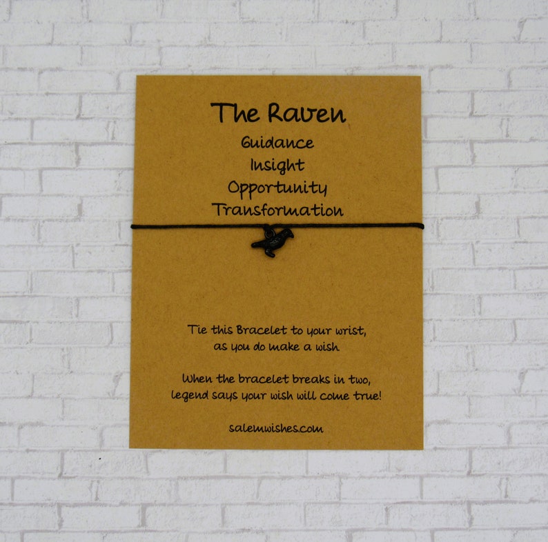 May include: A brown card with the words "The Raven" and "Guidance, Insight, Opportunity, Transformation" printed on it. A black raven charm is attached to a black string bracelet.