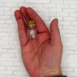May include: A small, clear glass bottle with a cork stopper. The bottle is about 1 inch tall and is held in a person's hand.