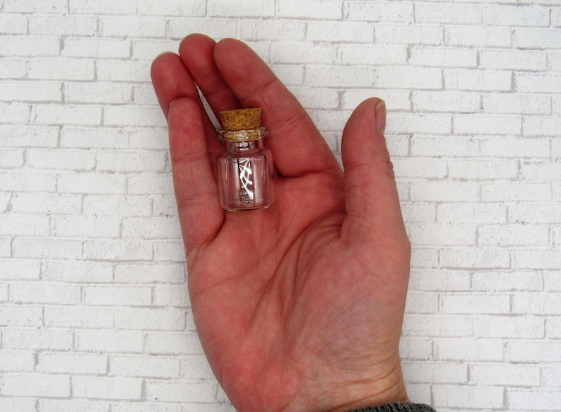 May include: A small, clear glass bottle with a cork stopper. The bottle is about 1 inch tall and is held in a person's hand.
