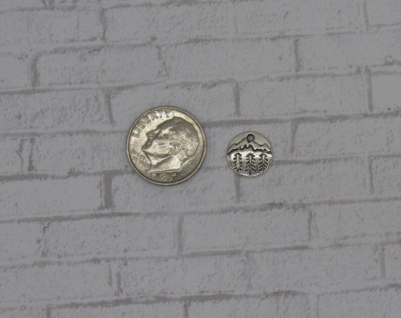 May include: Silver charm featuring a mountain and pine tree design. The charm is approximately the size of a ten pence coin.
