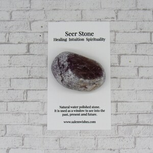 May include: A smooth, dark gray and white seer stone, also known as a water polished stone. The card reads "Seer Stone Healing Intuition Spirituality" and "Natural water polished stone. It is used as a window to see into the past, present and future. www.salemwishes.com"