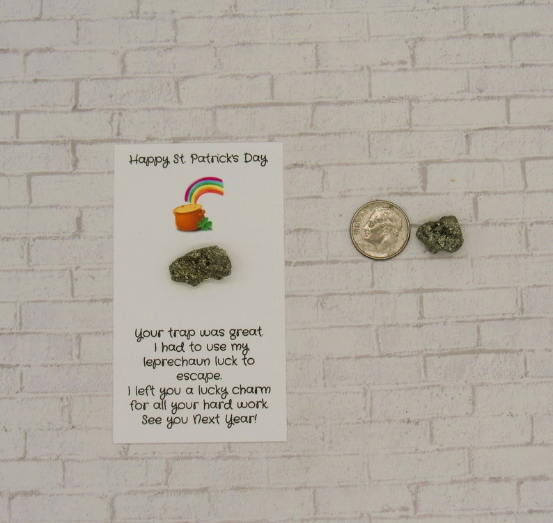 May include: A white card with a rainbow and a pot of gold illustration. The card reads "Happy St. Patrick's Day. Your trap was great. I had to use my leprechaun luck to escape. I left you a lucky charm for all your hard work. See you next year!" There are two small gold-colored rocks next to the card.