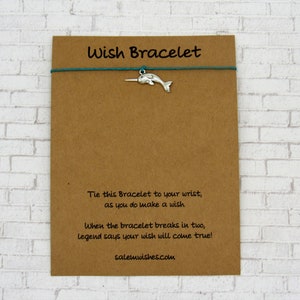 May include: A brown card with the text "Wish Bracelet" printed on it. The card has a teal string attached to it with a silver narwhal charm. The text on the card reads: "Tie this Bracelet to your wrist, as you do make a wish. When the bracelet breaks in two, legend says your wish will come true! salemwishes.com"