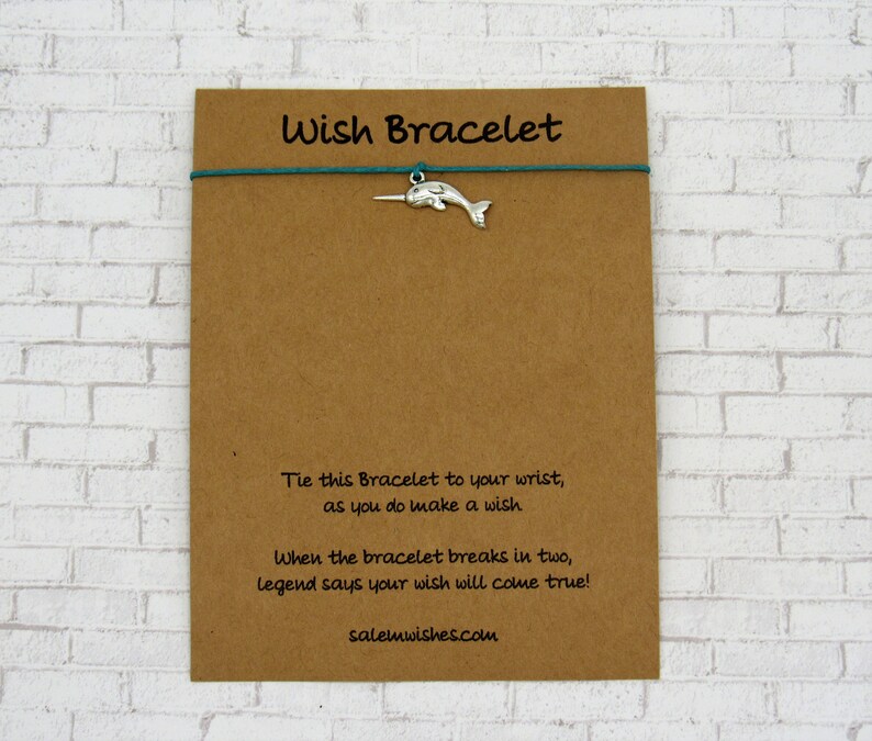 May include: A brown card with the text "Wish Bracelet" printed on it. The card has a teal string attached to it with a silver narwhal charm. The text on the card reads: "Tie this Bracelet to your wrist, as you do make a wish. When the bracelet breaks in two, legend says your wish will come true! salemwishes.com"