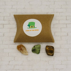 May include: A brown paper pouch with a white sticker that says "Luck of the Irish" and a green leprechaun hat with a four-leaf clover. Three crystals are in front of the pouch: a clear orange crystal, a green crystal, and a brown and gold crystal.