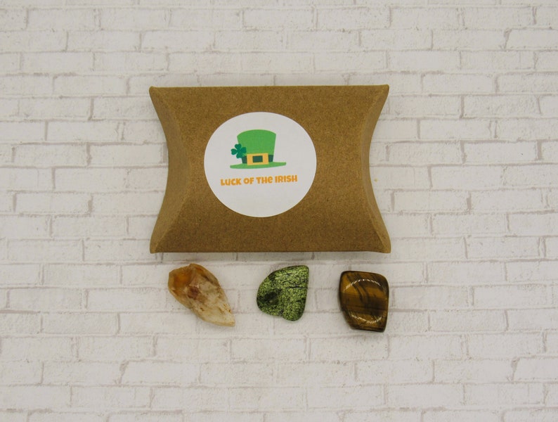 May include: A brown paper pouch with a white sticker that says "Luck of the Irish" and a green leprechaun hat with a four-leaf clover. Three crystals are in front of the pouch: a clear orange crystal, a green crystal, and a brown and gold crystal.