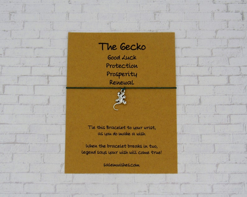 May include: A brown card with the text "The Gecko" and a list of words: "Good Luck", "Protection", "Prosperity", "Renewal". A green string bracelet with a silver gecko charm is attached to the card. The card also includes the text: "Tie this Bracelet to your wrist, as you do make a wish. When the bracelet breaks in two, legend says your wish will come true! salemwishes.com"