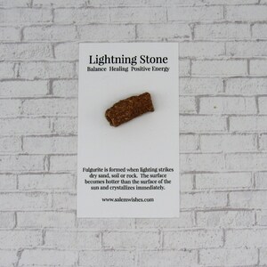 Pode incluir: Uma pedra de raio de fulgurite em um cart&atilde;o branco com texto. A pedra tem uma forma cil&iacute;ndrica &aacute;spera com uma superf&iacute;cie marrom e texturizada. O cart&atilde;o diz "Lightning Stone" e descreve sua forma&ccedil;&atilde;o.