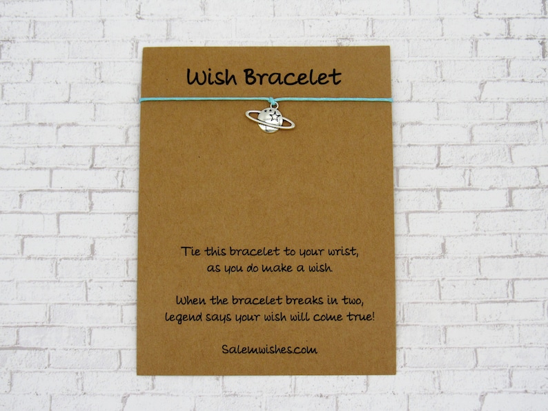 May include: A brown card with the text "Wish Bracelet" printed on it. A light blue string bracelet with a silver charm of a planet with a ring around it is attached to the card. The text on the card reads: "Tie this bracelet to your wrist, as you do make a wish. When the bracelet breaks in two, legend says your wish will come true! Salemwishes.com"