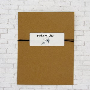 May include: A thin black cord bracelet attached to a rectangular white card. The card features the text "Make A Wish" in black lettering above a graphic illustration of two dandelion seed heads with seeds blowing in the wind on a brown background.