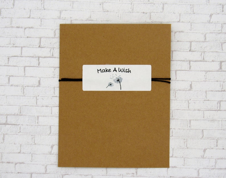 May include: A thin black cord bracelet attached to a rectangular white card. The card features the text "Make A Wish" in black lettering above a graphic illustration of two dandelion seed heads with seeds blowing in the wind on a brown background.