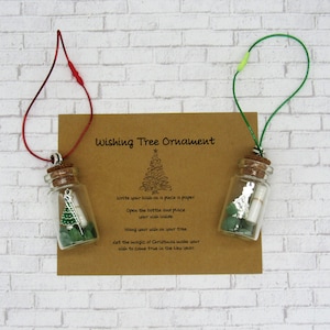 May include: Two glass wishing tree ornaments with cork stoppers and green string. Each ornament contains a small rolled piece of paper and green stones. The ornaments are hanging from a brown card with the text "Wishing Tree Ornament" and instructions on how to use the ornaments.