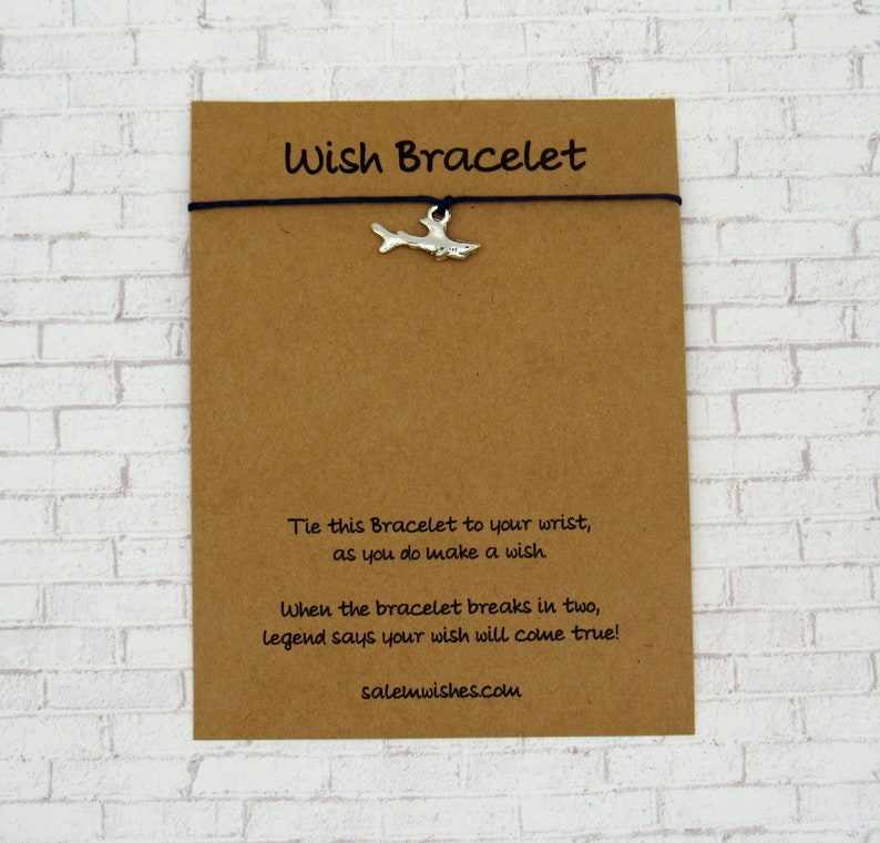 May include: A brown card with the text "Wish Bracelet" printed on it. The card has a black string with a silver shark charm attached to it. The text on the card reads: "Tie this Bracelet to your wrist, as you do make a wish. When the bracelet breaks in two, legend says your wish will come true! salemwishes.com"