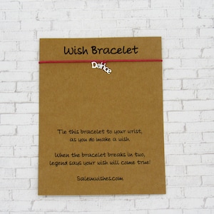 May include: A red string bracelet with a silver charm that says "Dafice". The bracelet is on a brown card with the text "Wish Bracelet" and instructions on how to make a wish.