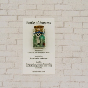 May include: A small glass bottle filled with green, blue, and white stones and a silver key charm. The bottle has a blue and white cork stopper. The label reads "Bottle of Success" and lists the stones as Amazonite, Aventurine, and Sodalite, with their meanings.