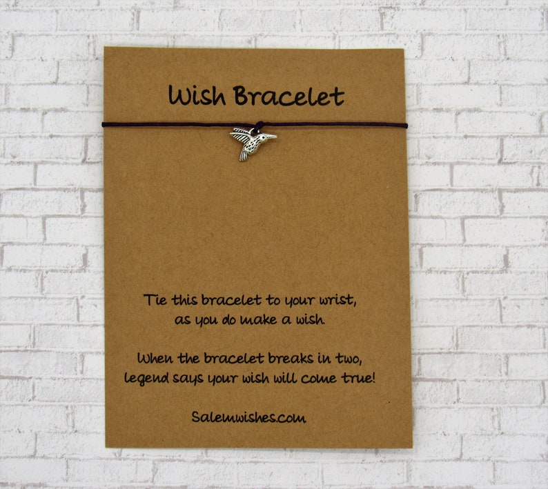 May include: A brown card with the text "Wish Bracelet" printed on it. A brown string bracelet with a silver hummingbird charm is attached to the card. The text on the card reads: "Tie this bracelet to your wrist, as you do make a wish. When the bracelet breaks in two, legend says your wish will come true! Salemwishes.com"