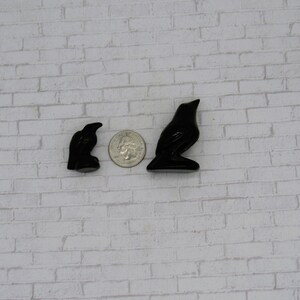 Pode incluir: Duas estatuetas de corvo pretas, uma pequena e outra maior, s&atilde;o exibidas ao lado de uma moeda para compara&ccedil;&atilde;o de tamanho. As estatuetas de corvo s&atilde;o brilhantes e t&ecirc;m um acabamento liso. O fundo &eacute; uma parede de tijolos brancos.