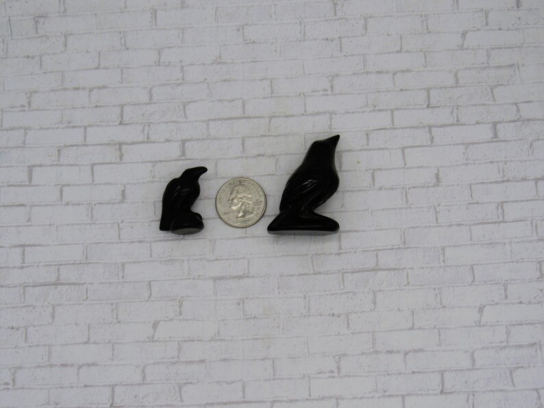 Pode incluir: Duas estatuetas de corvo pretas, uma pequena e outra maior, s&atilde;o exibidas ao lado de uma moeda para compara&ccedil;&atilde;o de tamanho. As estatuetas de corvo s&atilde;o brilhantes e t&ecirc;m um acabamento liso. O fundo &eacute; uma parede de tijolos brancos.