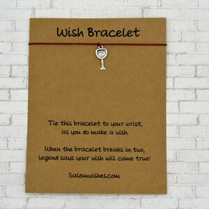 May include: A wish bracelet on a brown card. The bracelet has a red string and a silver wine glass charm. Text on the card reads "Wish Bracelet" and instructions on how to use the bracelet. The website Salemwishes.com is also printed on the card.