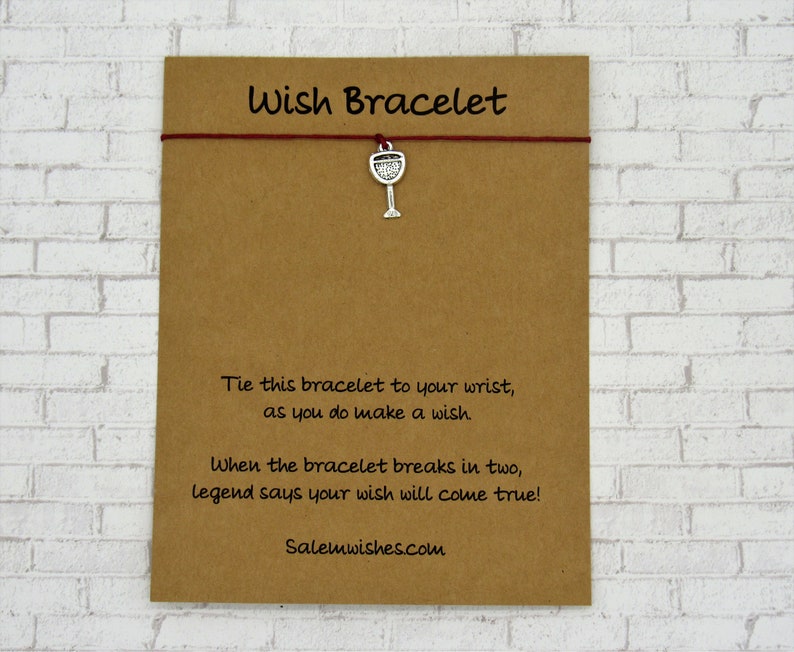 May include: A wish bracelet on a brown card. The bracelet has a red string and a silver wine glass charm. Text on the card reads "Wish Bracelet" and instructions on how to use the bracelet. The website Salemwishes.com is also printed on the card.