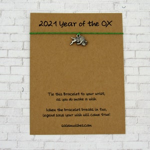May include: A brown card with the text "2021 Year of the OX" and a green string with a silver ox charm. The card also includes the text "Tie this Bracelet to your wrist, as you do make a wish. When the bracelet breaks in two, legend says your wish will come true! salemwishes.com".