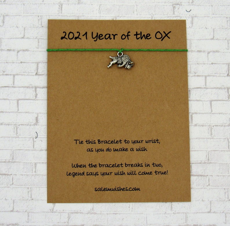May include: A brown card with the text "2021 Year of the OX" and a green string with a silver ox charm. The card also includes the text "Tie this Bracelet to your wrist, as you do make a wish. When the bracelet breaks in two, legend says your wish will come true! salemwishes.com".