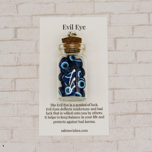 May include: A small clear glass bottle with a cork stopper filled with blue and white evil eye beads. The bottle is attached to a white display card featuring the text Evil Eye and a description of the symbol, ending with salemwishes.com.