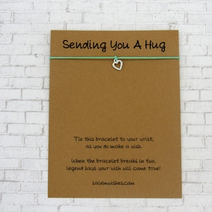 May include: A light green string bracelet with a silver heart charm. The card reads "Sending You A Hug" and includes instructions on how to tie the bracelet and make a wish. The card also includes the website salemwishes.com.
